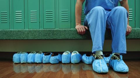 A hospital worker in PPE next to many pairs of shoes covered with hygiene show covers