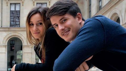 Young couple, one woman with dark long hair and a young man, with short dark brown hair and a moustache leaning over towards the camera. In the background there are stone buildings.