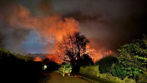 A street dimly lit by street lights. On the road are four fire officers. Behind them is a large orange fire and orange and grey smoke.