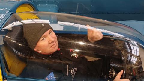 Dave Warby is sitting in the cockpit of the hydroplane Bluebird K7 with his thumbs up. Warby is wearing a grey hat and a black jacket with an Australian flag patch on the arm. He has one hand on the steering wheel and is covered by the clear pane over the blue water vehicle.