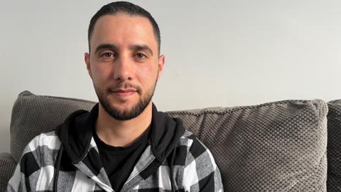 A man with dark, short hair looking into the camera with a small smile. He's sitting on a grey sofa and wearing a black and white checked hoodie.