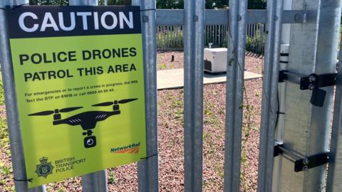 A sign is attached to a metal fence. It reads 'Caution. Police drones patrol this area." In the background, there is a white box containing the drone on an area of gravel.