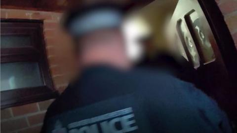 A still from police bodycam showing an unidentified policeman outside the front entrance to a home with an unidentified person answering the door during an arrest