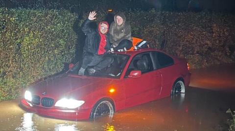 Davie Woodley-Kingston and his fiance Ashley Stevens are on the roof of a red BMW that is stuck in flood water. Davie is waving at the camera.