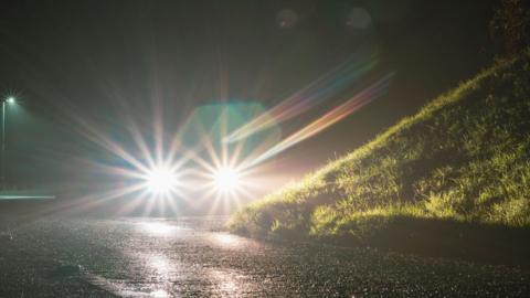 A generic picture showing a car's headlights glowing at night on an empty country road, with a grassy bank on the right.