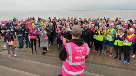 A large group of participants wearing bright running and support vests gather near a waterfront. The crowd appears engaged, many holding phones or clapping, creating an energetic and communal atmosphere.