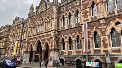 The exterior of the Royal Albert Memorial Museum in Exeter, which is in the style of Victorian Gothic Revival. The museum's exterior uses sandstone, has detailed stonework and uses characteristic Victorian features, including arched windows and ornate gables. People are walking past