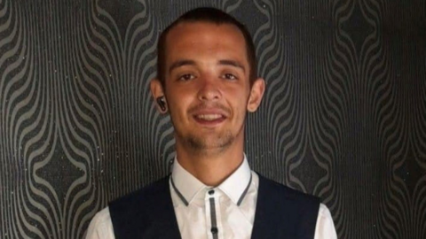 A man with short black hair is wearing a white shirt under a black waistcoat and is standing in front of a walk with black and silver wallaper