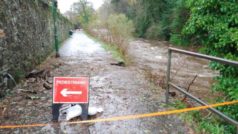 A closed of walkway in Tavistock. The pathway is wet from the overflowing river running alongside it on the right. There is a red road sign on the left which has the word "PEDESTRIANS" and a white arrow pointing left. There is a chunk of the pathway missing on the right.