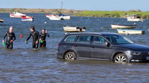 A dark blue VW estate car is surrounded by the sea and boats. Three people in wet suits are walking past the back of the car.