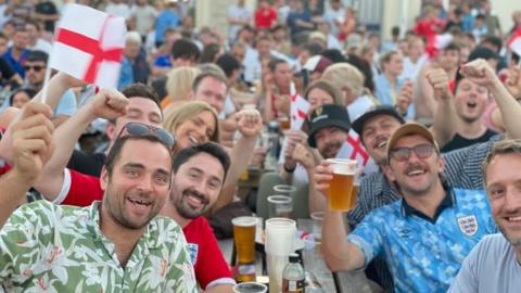 A crowd of people holding England flags and pints of beer are cheering and smiling