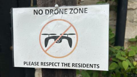 A white laminated piece of paper reads 'no drone zone, please respect the residents'. In the middle of the paper is a black drone graphic with a red cross in a circle over the top. The sign has been pinned to a wooden post outside a house.