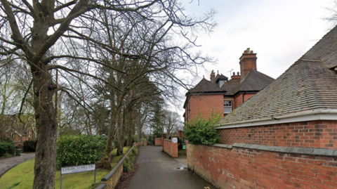 Buildings are visible on the right and far left of the photo. Trees, a bush and some grass are on the left and in the middle.