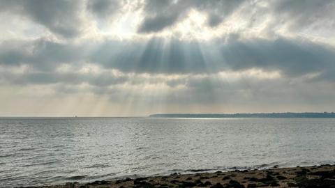 A beach scene is dominated by the sun's rays streaming through the clouds above. 