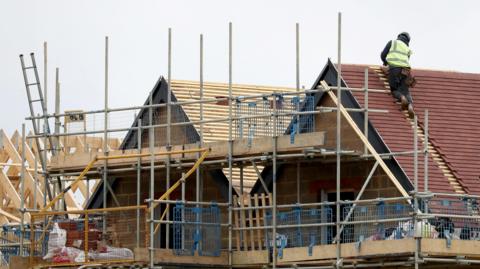 A construction worker on the roof of a house. It is on a building site, where several houses are being built. There is scaffolding in front of the buildings.