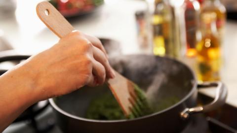 A person stirs a green substance in a black pot with a wooden spatula.  A kitchen counter and condiments are visible in the background but heavily blurred. 