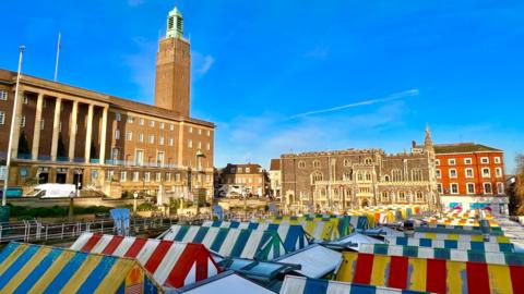 Rows of stalls at Norwich markets in Norwich city centre. The canopies are colourful and striped. In the background is the large imposing building of City Hall, with its tower furthest from the camera. There are blue skies above.