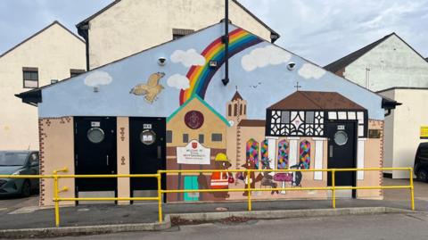 The front of a toilet block has been painted with images of the old black and white pillared town hall, St Bartholomew's church with stained glass windows and a Great Bustard flying in the sky. Cartoon animals can be seen in the foreground. And a large rainbow painted above.