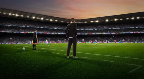 A promo image for Football Manager. A female manager stands on the sidelines of a football pitch. She's wearing white shoes, and a black suit with her hands in her pockets. It's evening time as the sun appears to be setting. The pitch is lit up, with players wearing red kits and white kits. A male referee stands further away with a flag in hand.