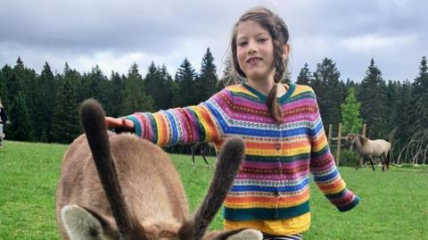 Aisha Cox smiles with a donkey. She is wearing a striped, multi coloured cardigan. In the background there is a forest lined with trees in a rural area.