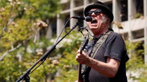 The rock star Neil Young plays a guitar and sings on a stage outside in the daytime. 