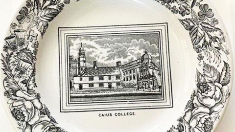 A white plate is decorated with a view of Caius Court in its centre, in black, and the words "Caius College" printed beneath. Around the edges are black drawings of flowers and foliage.
© Caius College