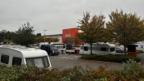 Several caravans parked in a car park outside a B&Q store in Patchway, near Bristol. There are trees and hedges in the car park and the orange storefront can be seen in the distance.