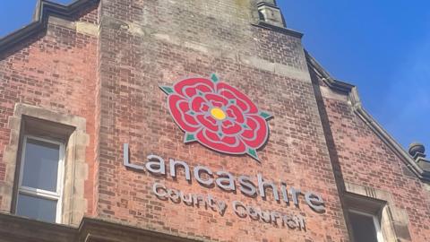 Lancashire County Council sign, with the red rose emblem, on the side of a tall, redbrick building. The photograph was taken on a sunny day with clear blue skies.