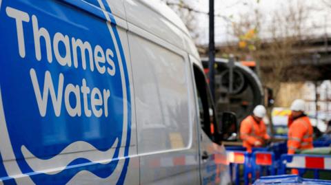 A white van with a blue Thames Water logo is parked in front of some blue construction barriers with white and orange reflective strips on them. There are two male construction workers, wearing orange hi-vis jackets, working behind the barriers. There is a railway bridge in the background.