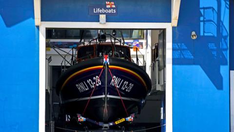 A blue building. There is a large hole in the front from where a lifeboat would launch. Seen through this hole we can see the dark blue hull of a lifeboat. A sign above the gap reads Lifeboats