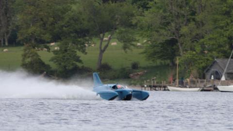 Bluebird K7 "planes" on Coniston Water. The blue craft is throwing up a large spray behind it as it glides on top of the lake's surface. On the far shore, sheep are grazing on an incline.