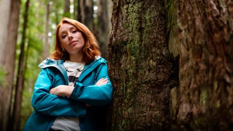 Dr Hannah Fry leaning up against a tree in a forrest.