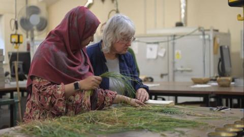 Two women are inside lab sorting through grass cuttings on a table in front of them while consulting an open book