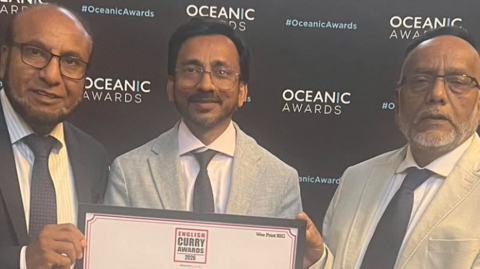 Three men wearing glasses, a shirt, a tie and a jacket are holding a framed certificate that includes the words outstanding restaurant of the year Basmati restaurant. They are in front of a black board including the words Oceanic Awards.