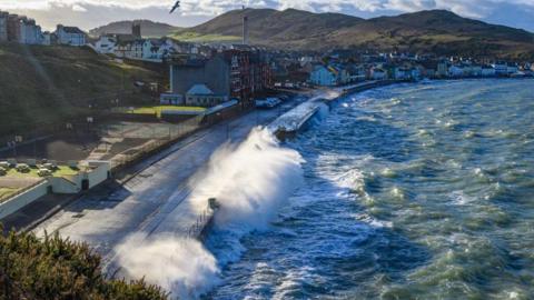 Peel Bay on a blustery day, the sun is shining. It is high tide and the waves crash off the harbour side. Rows of houses line the promenade. You can also see empty tennis courts next to them. There are rolling green hills in the background.