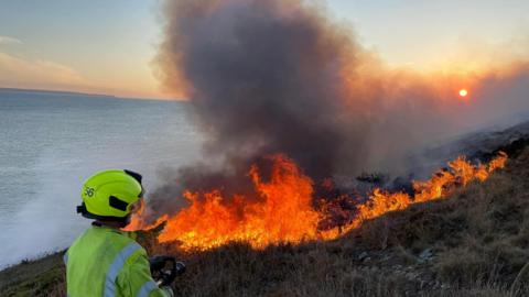 A firefighter in protective clothing standing at the edge of a moorland wildfire by the sea and using a swatter to try and extinguish the flames. Gorse and vegetation is ablaze in the background.
