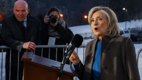 Hillary Clinton speaks to the press outside. She is wearing a blue top and a brown jacket