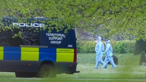 Two men in hazmat suits walk across the grass, shrouded by the branches of a tree. They walk towards a police van parked nearby.