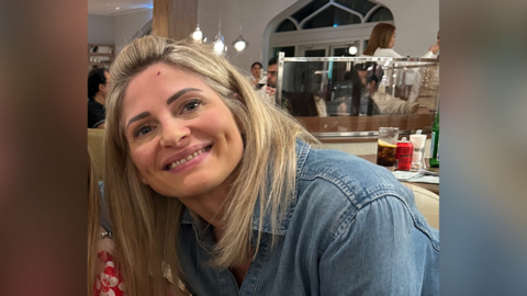 Ms Phipps is smiling at the camera and leaning to the left to pose with her daughter, who is just out of shot. She is in a restaurant and other diners can been seen in the background. Ms Phipps has shoulder length blonde hair and is wearing a blue denim shirt. 