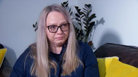 Magda Szmeichel has blond hair and black glasses. She sits on a sofa with yellow cusions and is visibly upset.