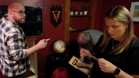 People inside a red room appearing to try to solve puzzles - a woman in a black top with blond hair on the right reads a note, a bald man on the left in a cheque short points at something on the wall, behind them a man in grey in bending down to look at something