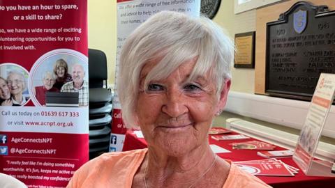 Janette, who is claiming pension credit, she is wearing a peach coloured top and is sitting in front of signs about Age Connects. She is looking at the camera.