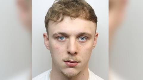 A custody mugshot of Braden Lynham. He has a small moustache and goatee beard, short curly light brown hair and is wearing a white T-shirt
