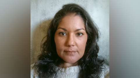 Ruth Bushi is looking straight into the camera while taking a selfie. She has long black hair and is wearing a grey jumper.