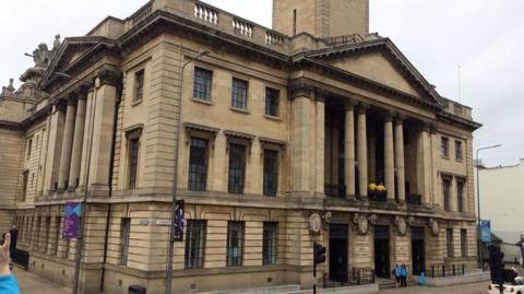 The Guildhall is made of stone and has four large pillars that hold up an apex roof above a balcony on the first floor.  It is a grand building with large black windows.