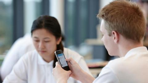 Boy dressed in school uniform with mousy sat in classroom looking at his phone. In the background, a girl with dark hair is looking down at her work.