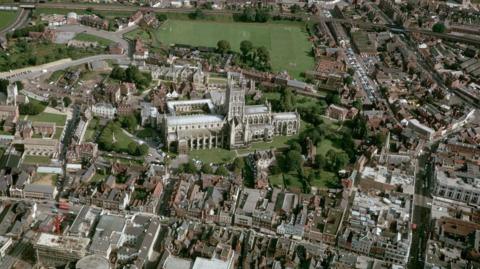 Aerial view of Gloucester, with cathedral in centre and green fields surrounding along with homes and roads.