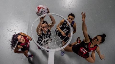 Shot from above of a netball hoop with four female players beneath. The woman second from left is about to put a white ball into the hoop. Furthest right (Farah Hasan) is sticking out her arm to try to block the ball. The others are looking at the hoop. There are all wearing dark blue and red netball vests and shorts. 