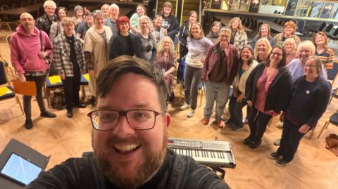 A wide-angle selfie taken indoors shows a large group of approximately two dozen people standing in rows on a light wooden floor, with rows of empty chairs behind them. In the foreground, one person holds the camera close to the lens, partially visible above a keyboard on a stand; behind the group are mirrors, black-and-gold decorative panels, and a staircase, suggesting a rehearsal or performance space.