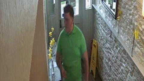 A white man is wearing a green polo shirt and is walking in a narrow hallway. Behind him is a door and a wet floor sign.